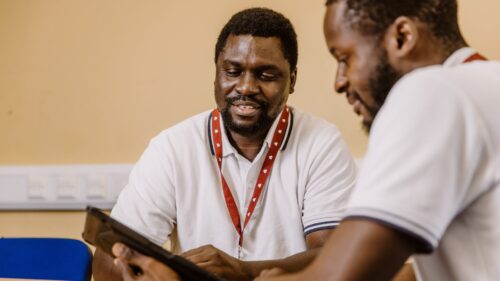 Two people wearing lanyards sit at a table, with one holding a tablet during a discussion.