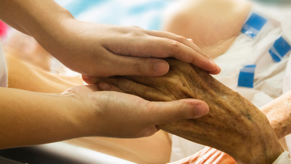 A pair of hands gently holds the hand of an older adult lying in bed.
