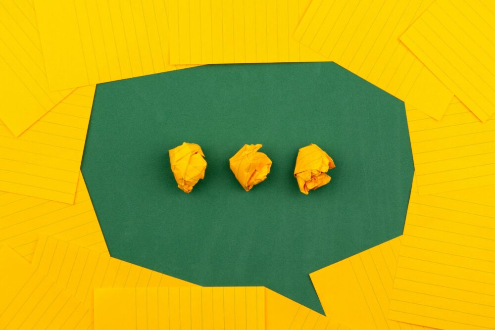 A green speech‑bubble shape with three crumpled yellow paper balls inside, surrounded by yellow notepaper.