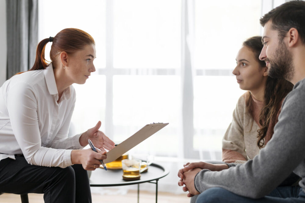 A person holding a clipboard speaks with two people seated together during a conversation.