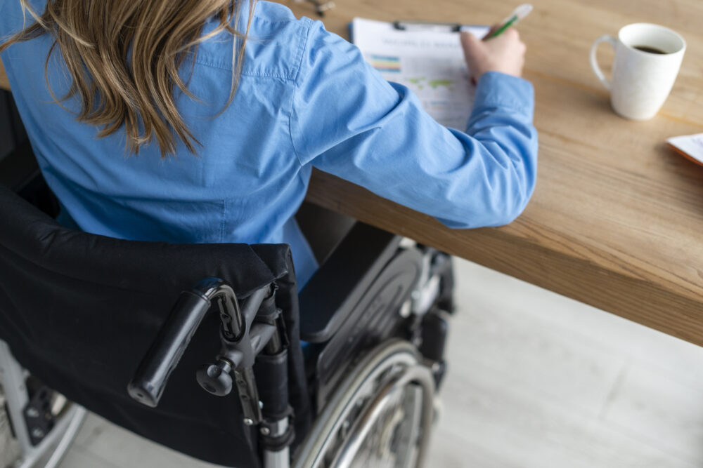 A person in a wheelchair sits at a desk writing on a clipboard.