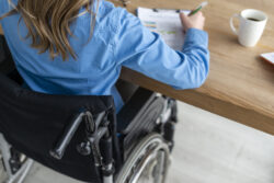 A person in a wheelchair sits at a desk writing on a clipboard.