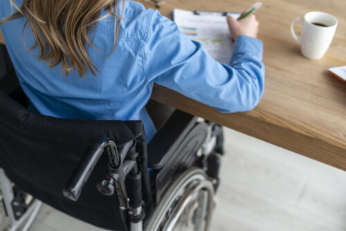 A person in a wheelchair sits at a desk writing on a clipboard.