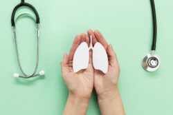 Hands hold a paper cut‑out of lungs on a green background beside two stethoscopes.
