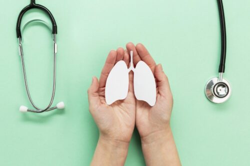 Hands hold a paper cut‑out of lungs on a green background beside two stethoscopes.