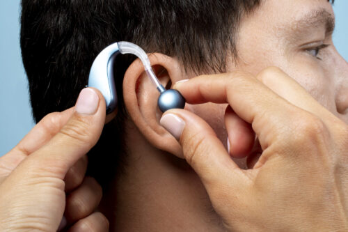 Hands position a behind‑the‑ear hearing aid onto someone’s ear.