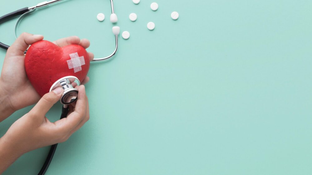 Hands hold a stethoscope against a red heart model with a small bandage on it, with tablets scattered nearby.