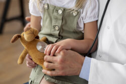 A child holds a stuffed animal while a healthcare professional places a stethoscope on it.