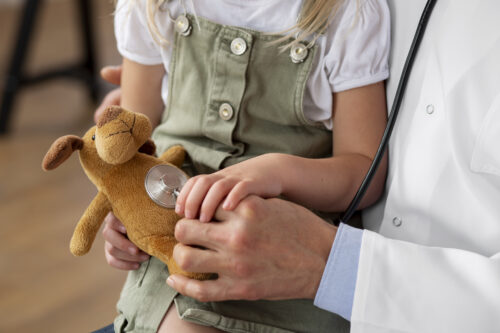 A child holds a stuffed animal while a healthcare professional places a stethoscope on it.