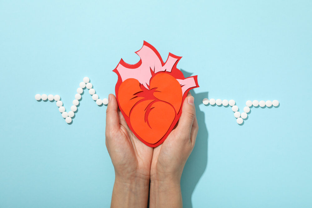Hands hold a paper heart shape in front of a light blue background with a heartbeat line made from small white tablets.