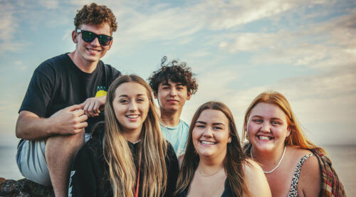 A group of five young people sit and stand together outdoors with the sky in the background.