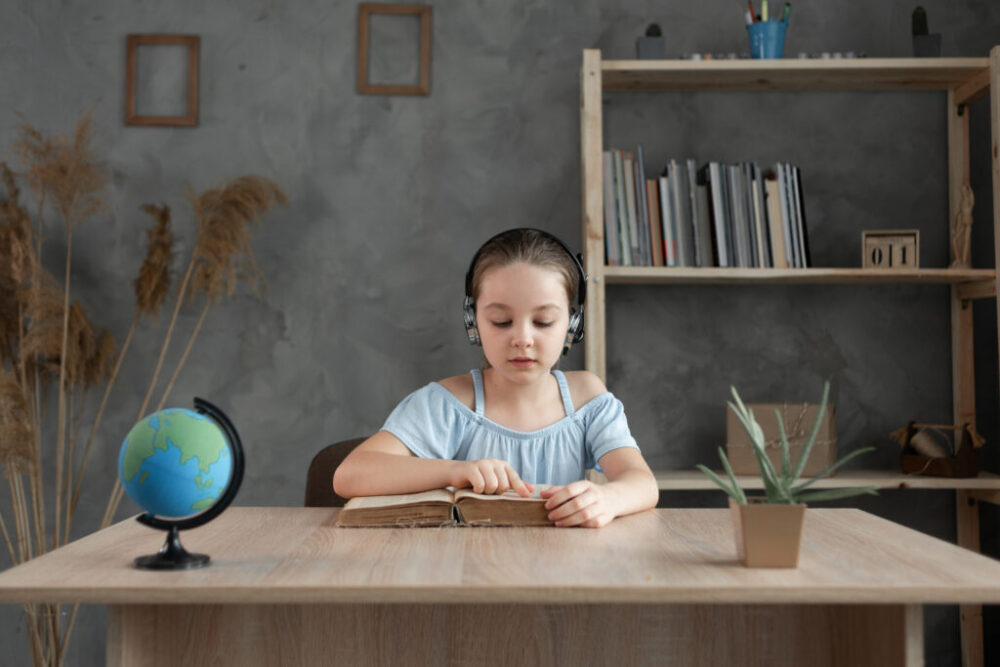 A child sits at a desk with headphones on, reading an open book, with shelves and a small globe in the background.