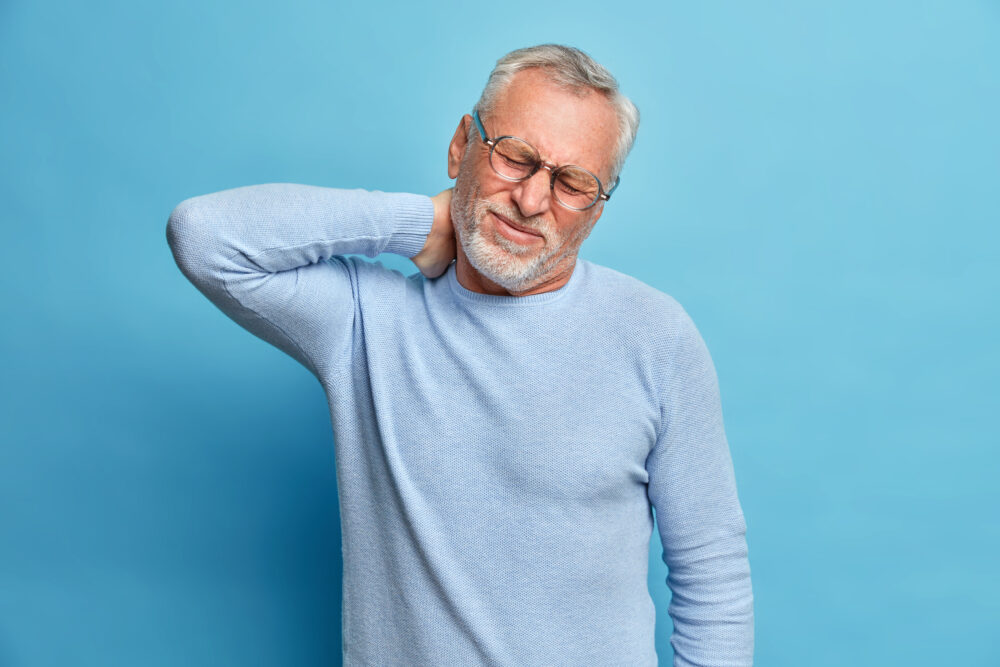 An older adult stands against a blue background with one hand on the back of the neck, suggesting possible discomfort.
