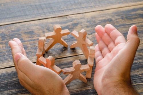 Two hands are cupped around a circle of small wooden human‑shaped figures on a wooden surface.