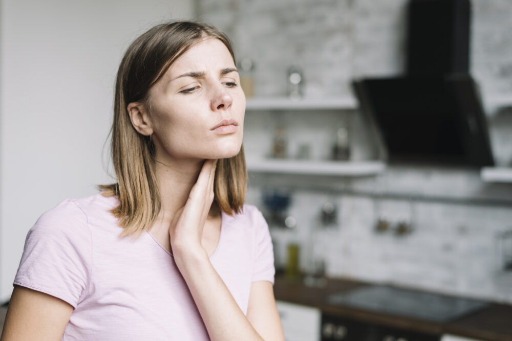 A person indoors touches the front of the neck with one hand, suggesting possible neck pain.