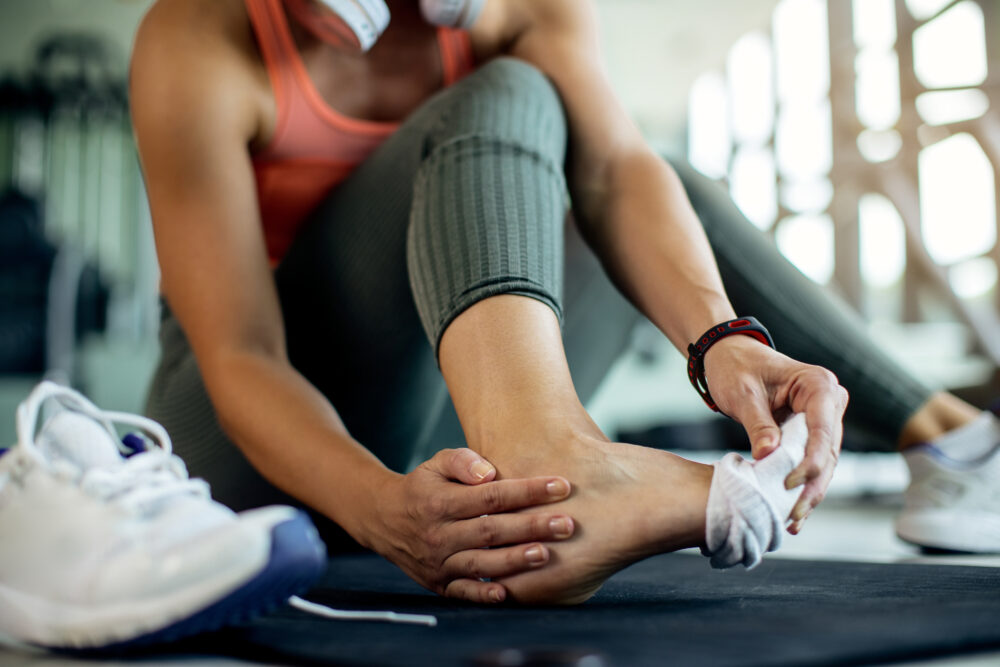A person sitting on a gym floor holds their foot with both hands, suggesting a possible foot problem.