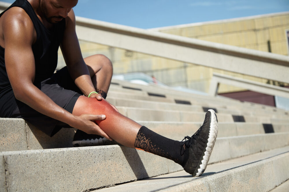 A person sits on outdoor steps holding a reddened lower leg, suggesting possible discomfort.
