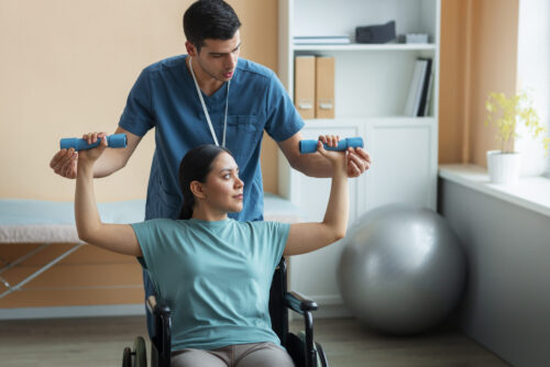 A healthcare professional assists a person in a wheelchair with lifting small dumbbells during an exercise session in a therapy room.