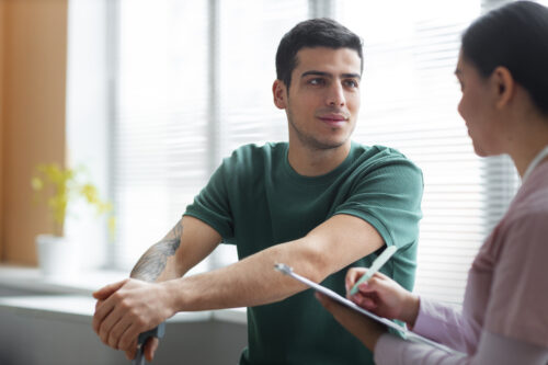 A person sits across from a healthcare professional who is holding a clipboard during a consultation in a bright room.