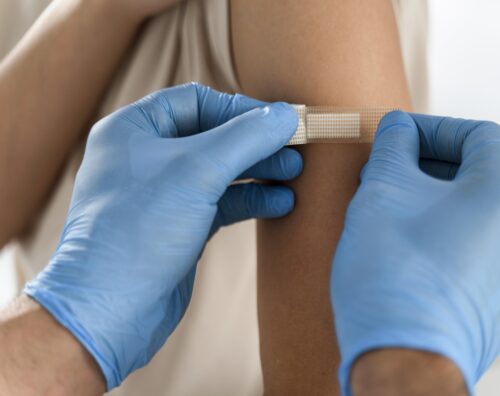 Gloved hands place a small adhesive bandage on someone’s upper arm.
