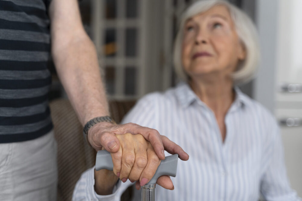 A seated older adult holds a walking stick while another person places a supportive hand over theirs.
