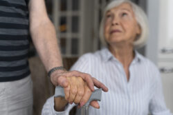 A seated older adult holds a walking stick while another person places a supportive hand over theirs.
