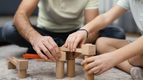 Hands build a structure using wooden blocks while sitting on the floor.