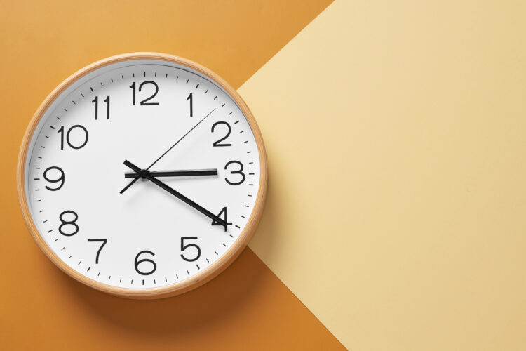 A round wall clock with a white face and black numbers hangs against a two‑tone orange and beige background.