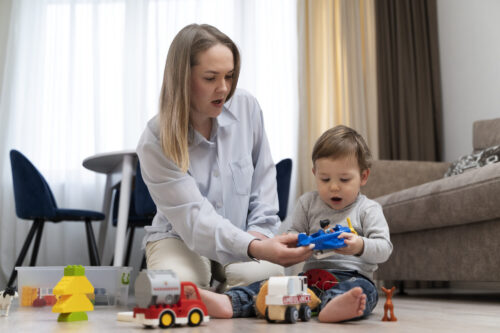 An adult sits on the floor with a child, helping the child play with toy vehicles in a living room.