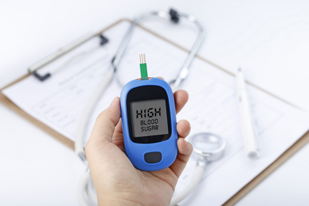 A hand holds a blood glucose meter displaying a high blood sugar reading, with medical tools on the desk behind.