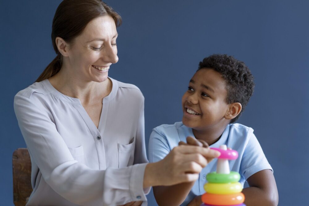 An adult and a child sit together at a table, placing colourful stacking rings on a toy.