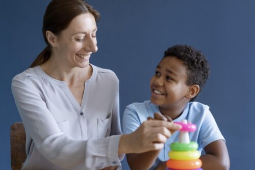 An adult and a child sit together at a table, placing colourful stacking rings on a toy.