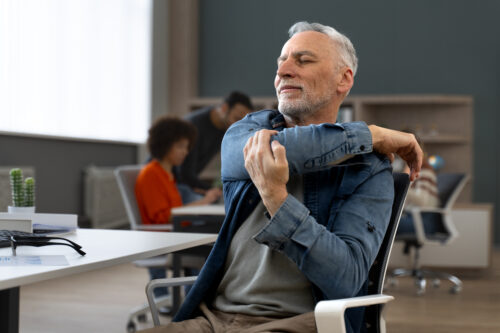 A person sitting in an office chair stretches an arm across the body, with the posture hinting at possible arm or shoulder discomfort.