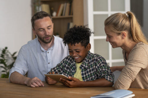 Two adult and a child sit at a table looking at a tablet together.