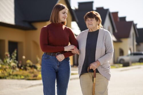 A person walks outdoors supporting an elderly person who is using a walking stick.