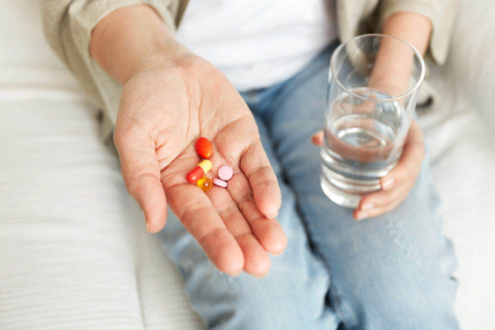 An open hand holds several colourful tablets and capsules, with a glass of water in the other hand.