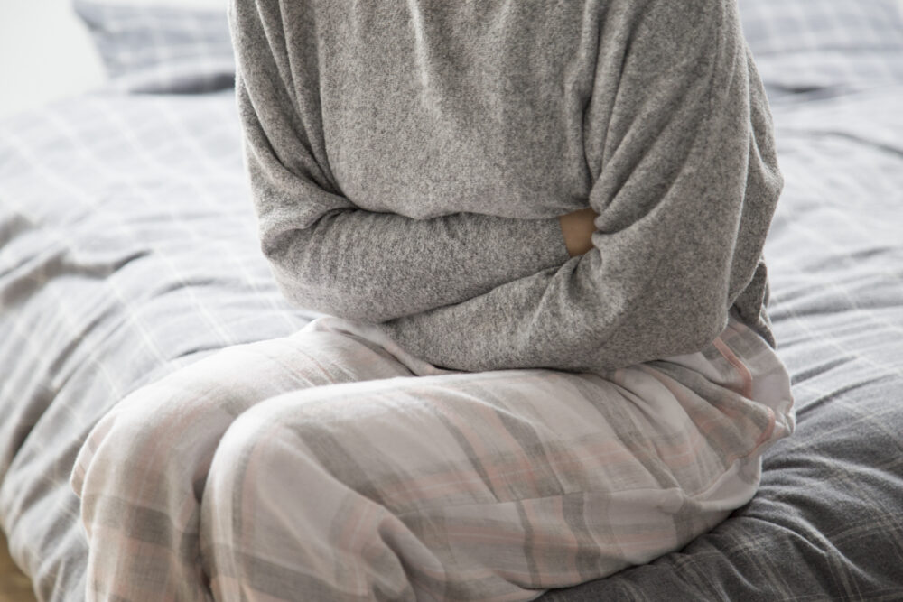 A person sits on a bed holding their stomach with both arms.