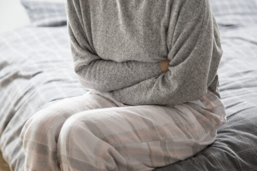 A person sits on a bed holding their stomach with both arms.