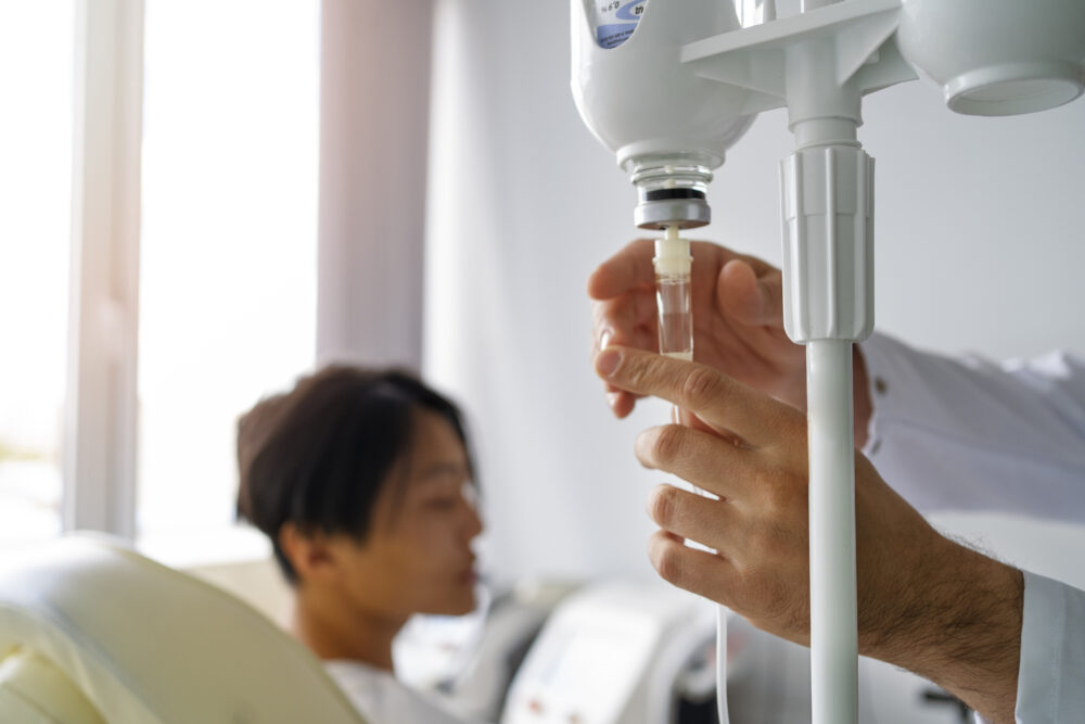 A medical professional adjusts an IV drip while a person sits nearby in a clinical setting.
