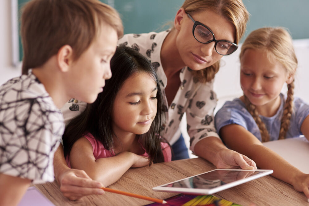 Adult helping children use a tablet at a table.